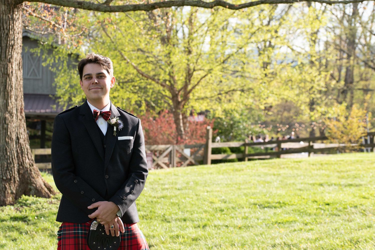 Groom in a black jacket and red tartan kilt stands on green grass beside a large tree at an outdoor wedding.