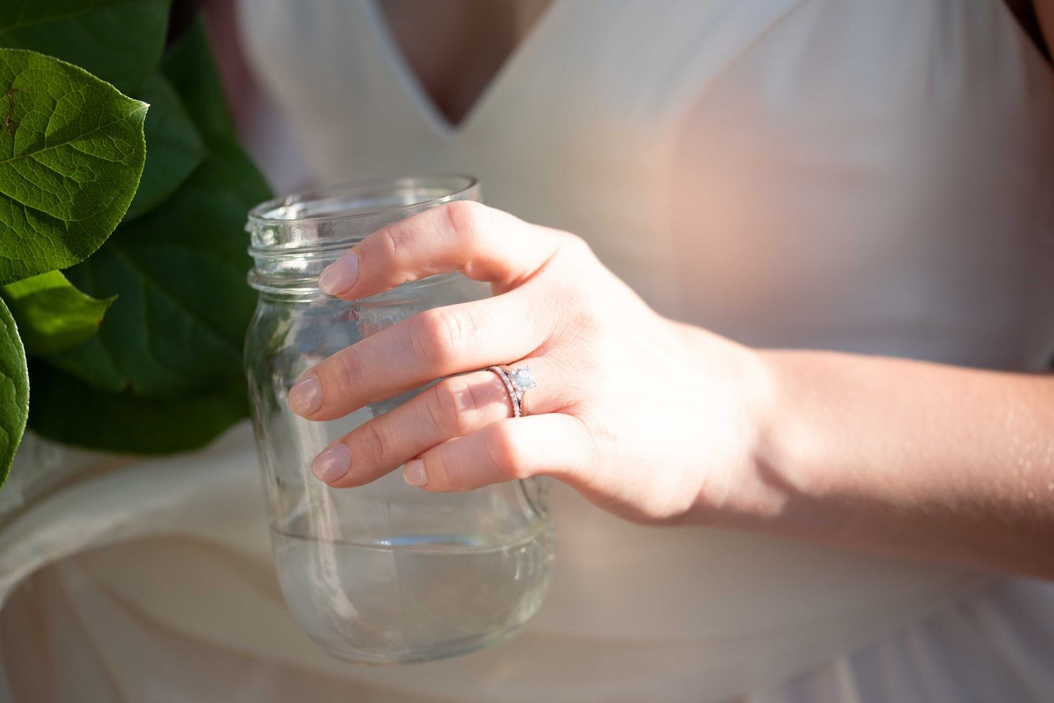 Bride wearing engagement ring holding a clear mason jar near green foliage.