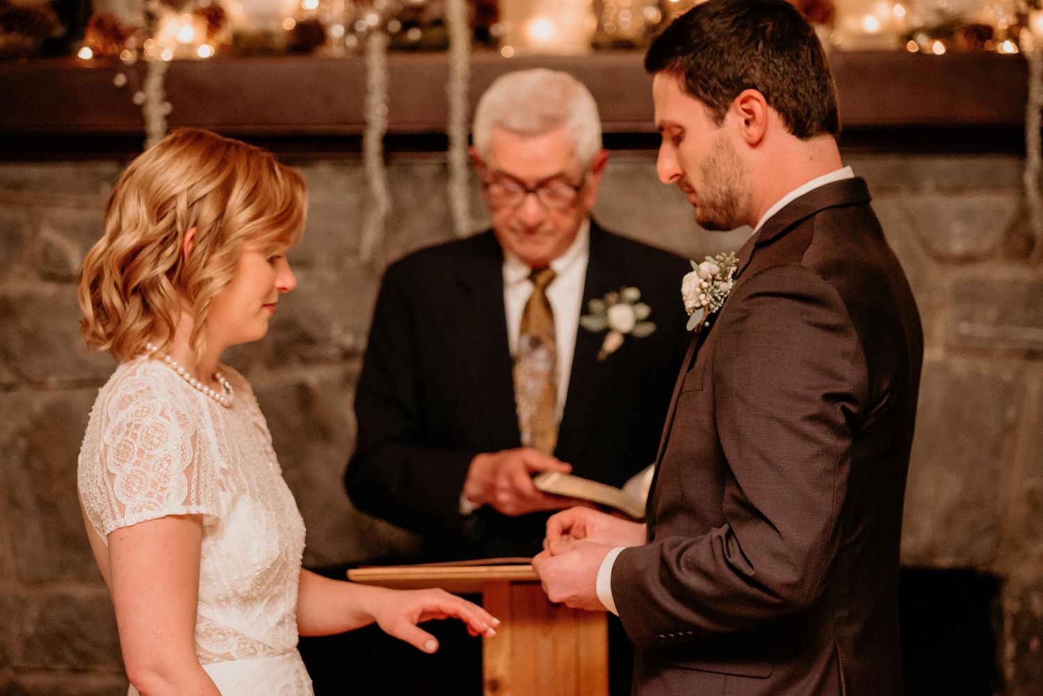 Bride and groom exchanging rings during wedding ceremony with officiant in background near stone fireplace.