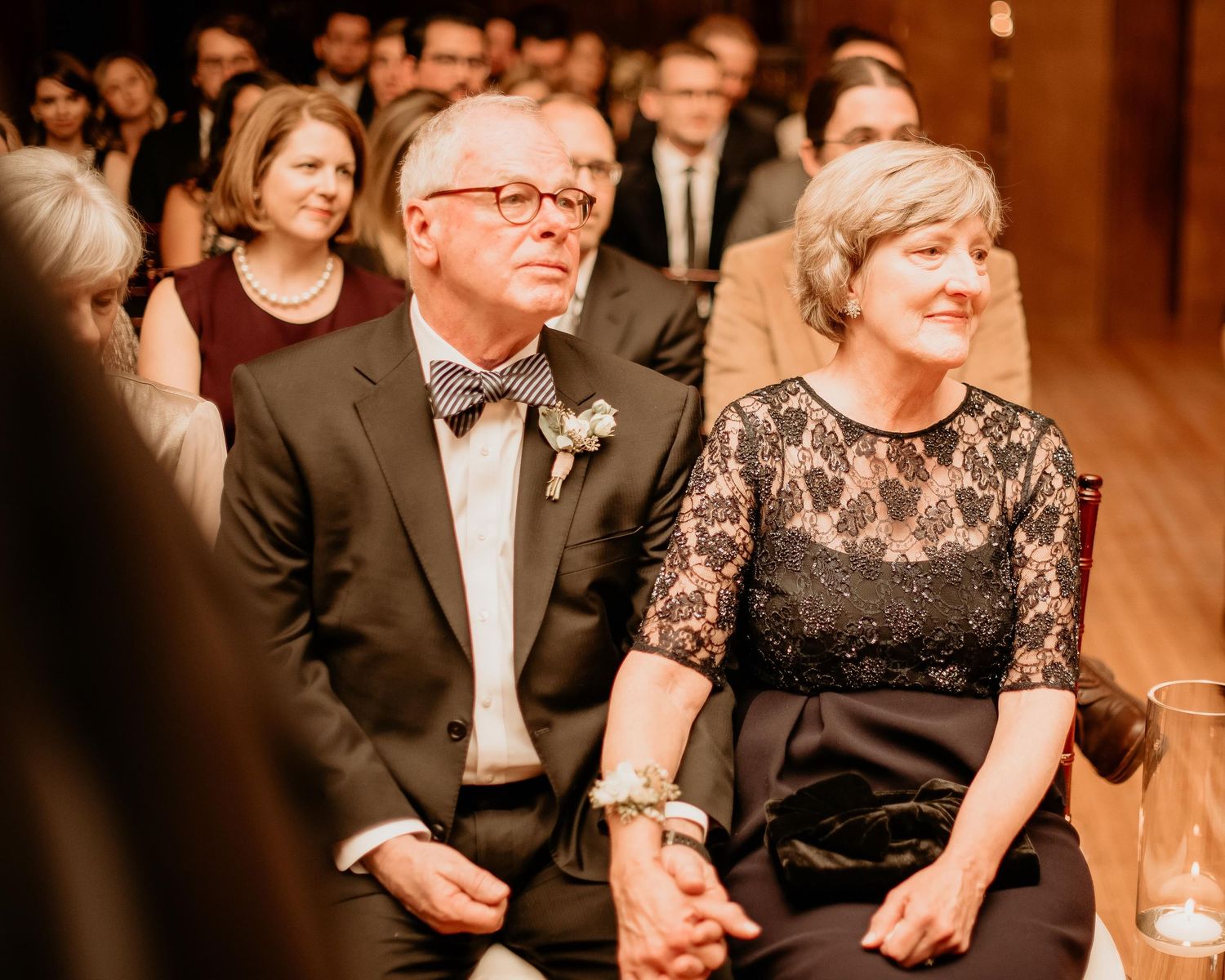 Elegant couple holding hands at a formal wedding ceremony, man in bow tie with boutonniere, woman in black lace dress.