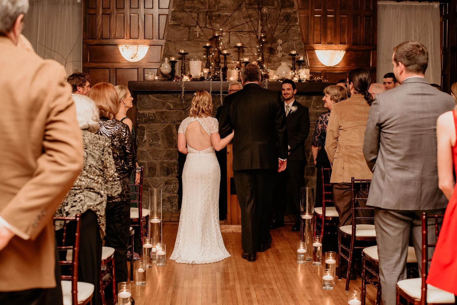 Elegant indoor wedding ceremony with bride and groom at candlelit altar, guests standing in aisle, stone fireplace backdrop.