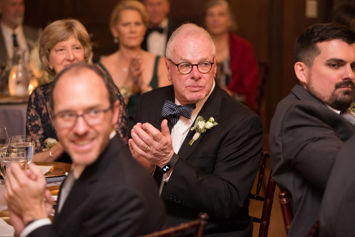 Guests applauding at a formal wedding reception, with an older man in a bow tie and boutonniere clapping in the foreground.