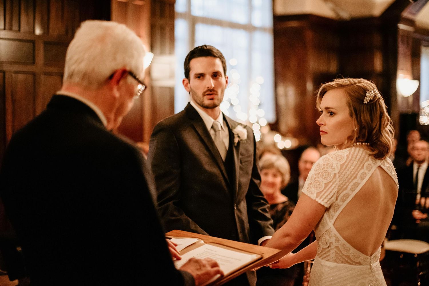 Bride and groom exchange vows at elegant wood-paneled wedding ceremony with officiant holding open book.