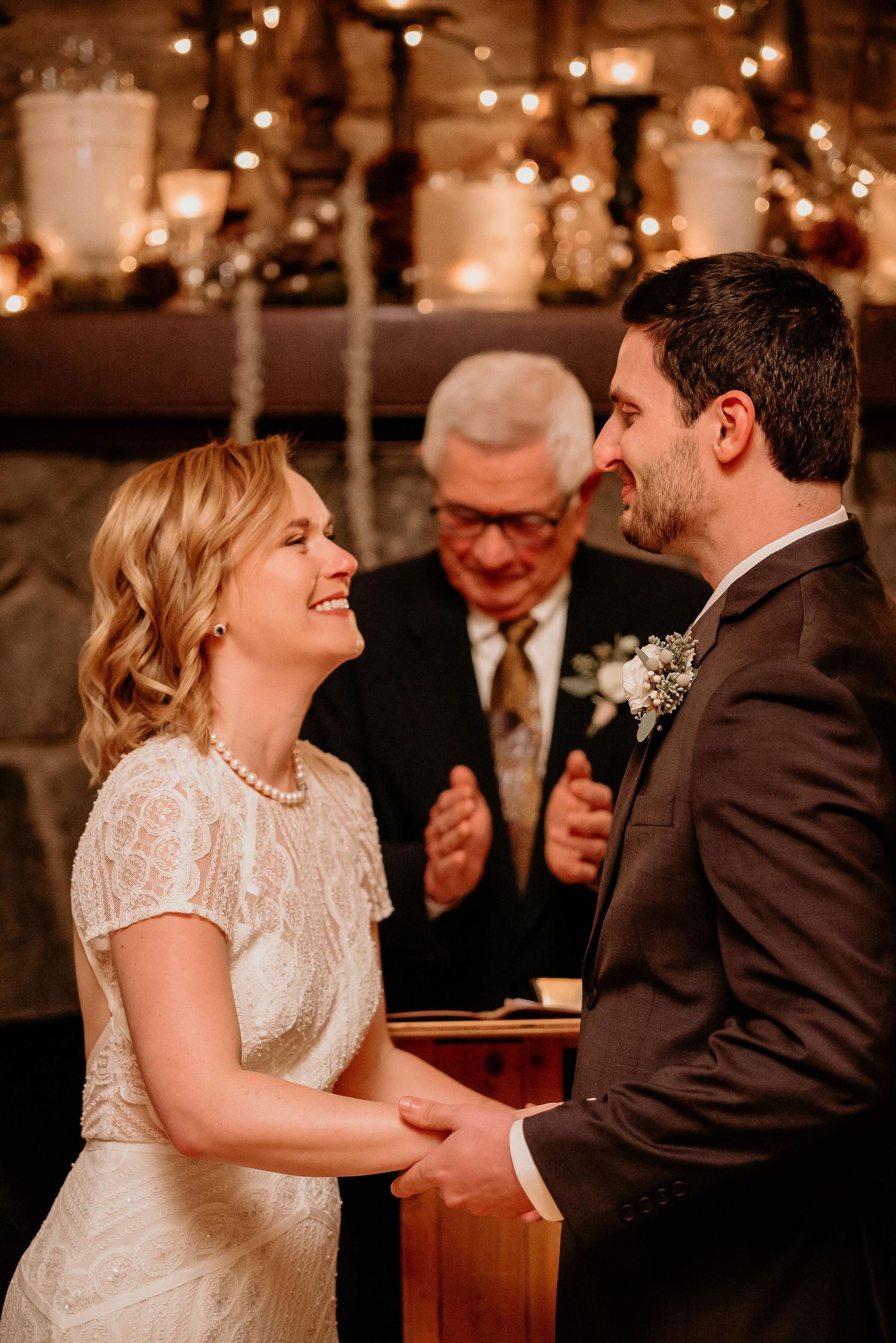 Bride and groom smiling at each other during wedding ceremony with candles and fairy lights in background.
