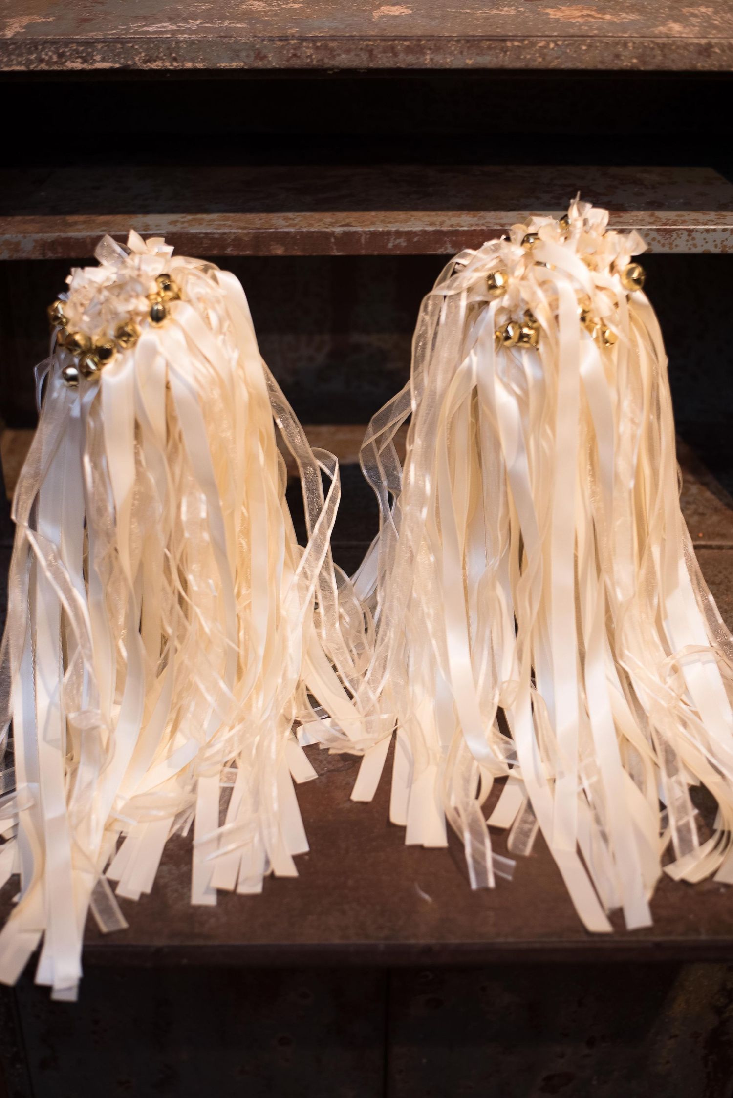 Two ivory ribbon wand pom-poms with gold jingle bells displayed on rustic wooden shelves.