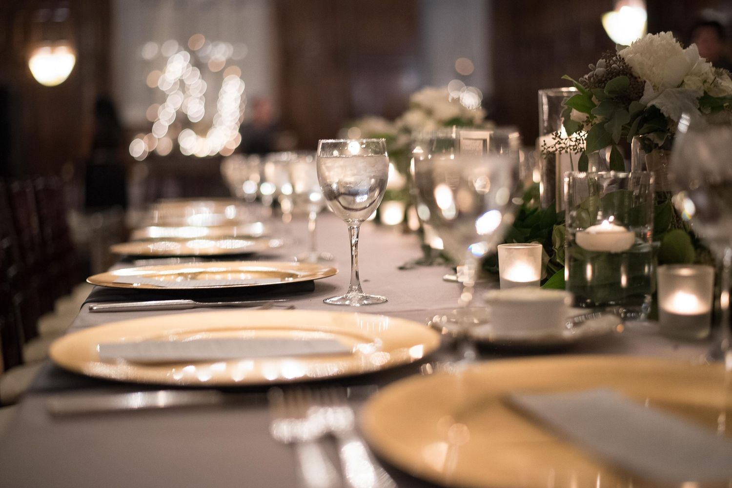 Elegant wedding reception table setting with gold charger plates, crystal glasses, and white floral centerpieces with candles.
