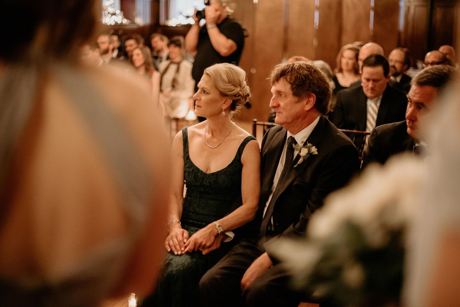 Couple seated together at a wedding ceremony in an elegant venue, dressed in formal attire, holding hands.