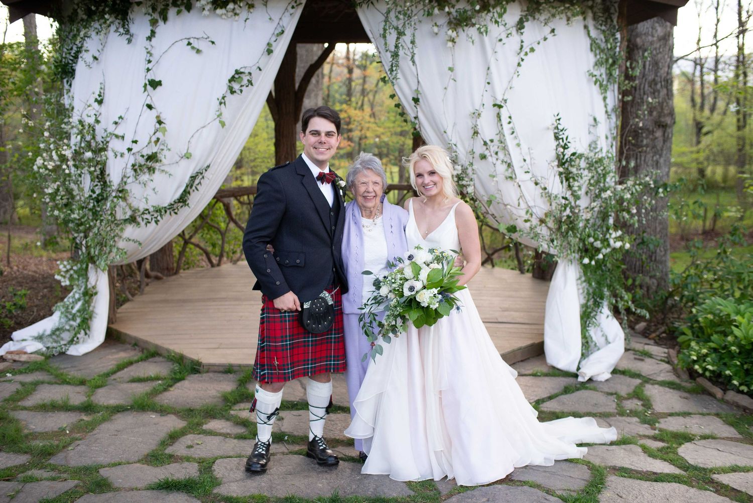 Bride and groom in red tartan kilt with elderly woman posing under floral wedding arch outdoors.