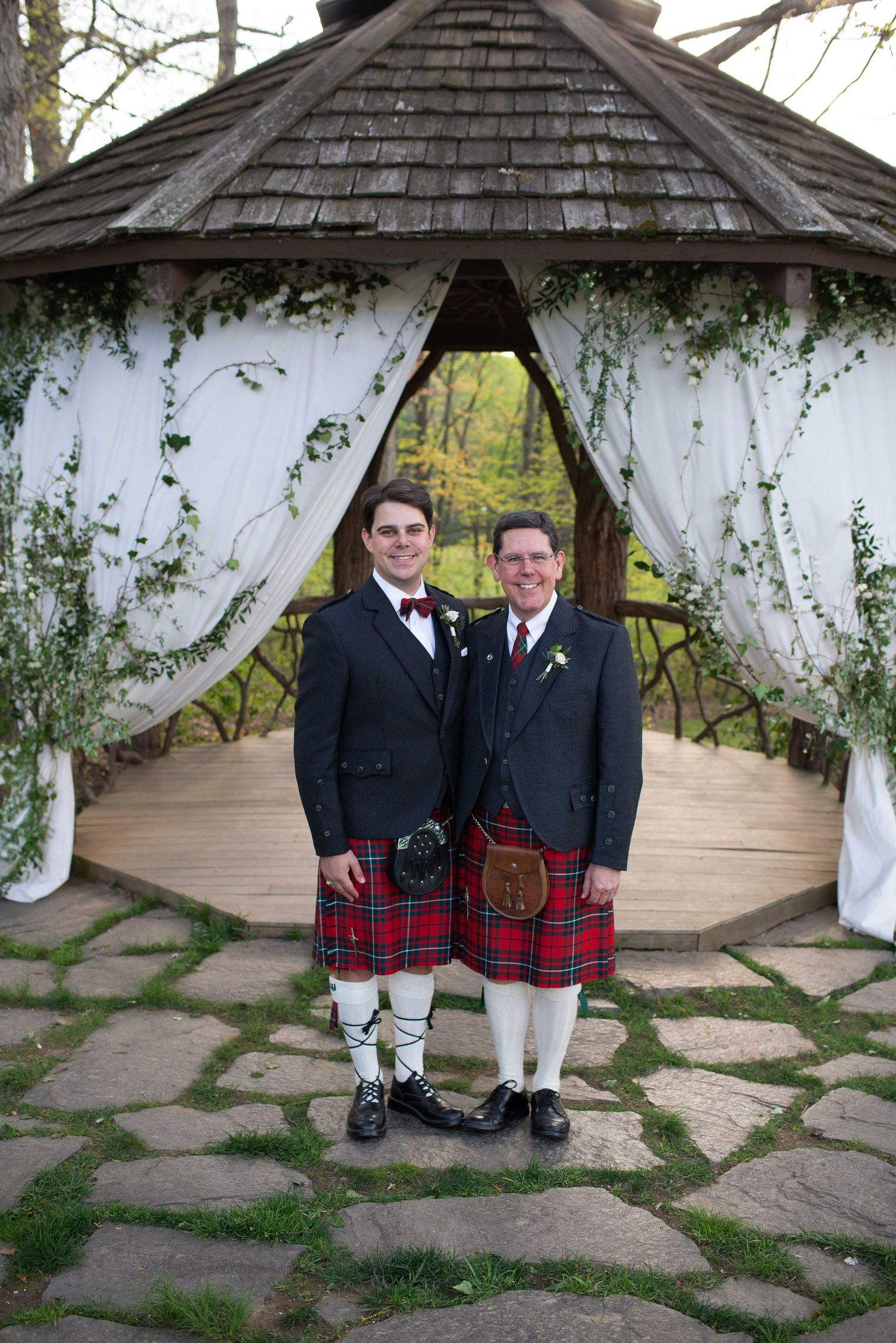 Two men in traditional Scottish kilts and formal jackets pose together under a decorated gazebo at an outdoor wedding.