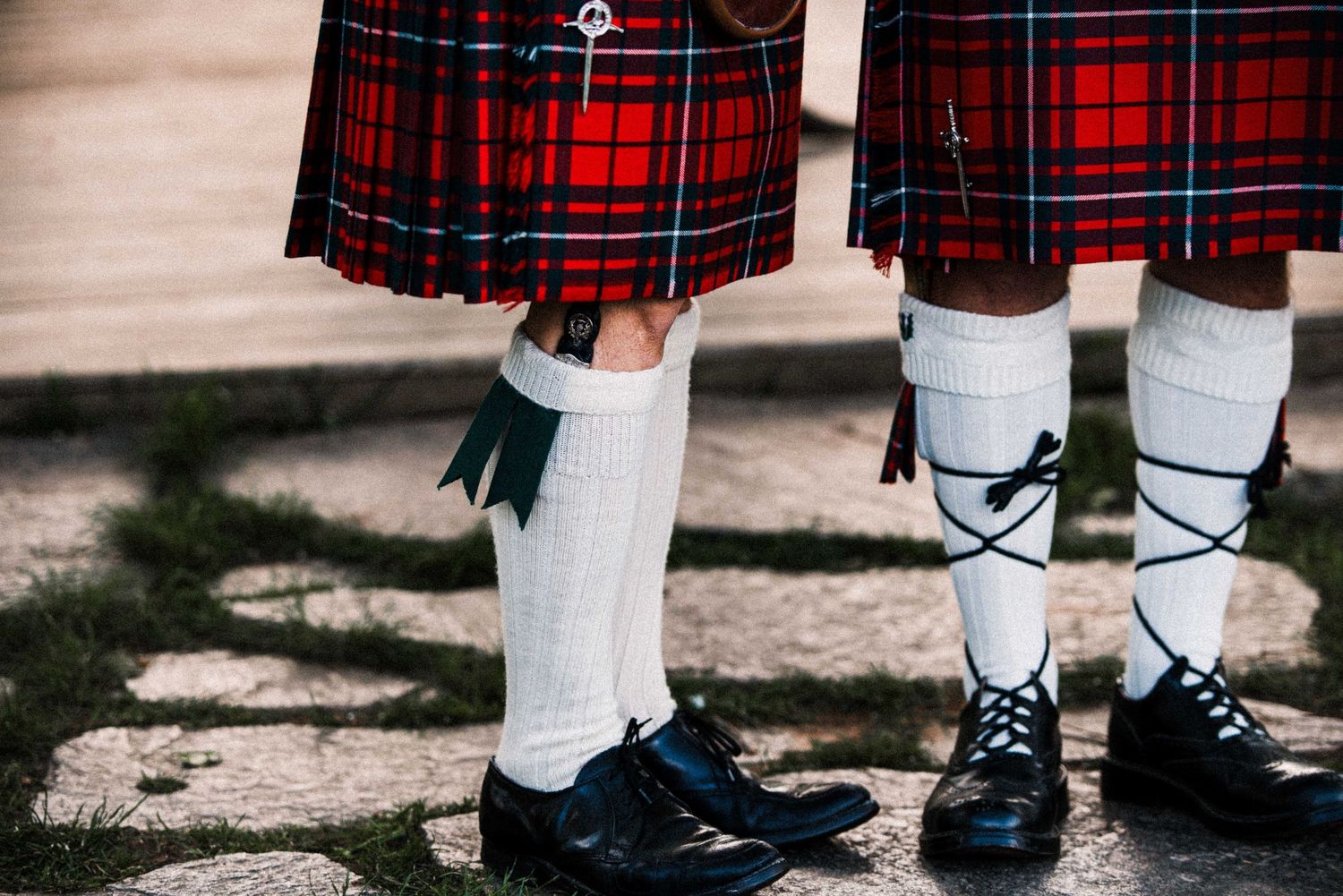 Two men wearing traditional Scottish red tartan kilts, white knee-high socks, and black dress shoes stand outdoors.