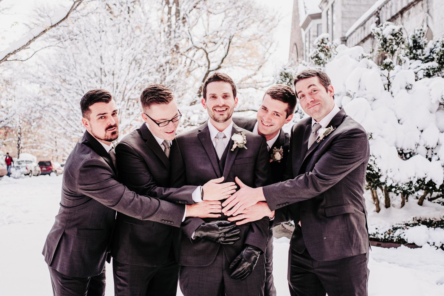 Five groomsmen in dark suits smiling and posing together outdoors in a snowy winter wedding setting.