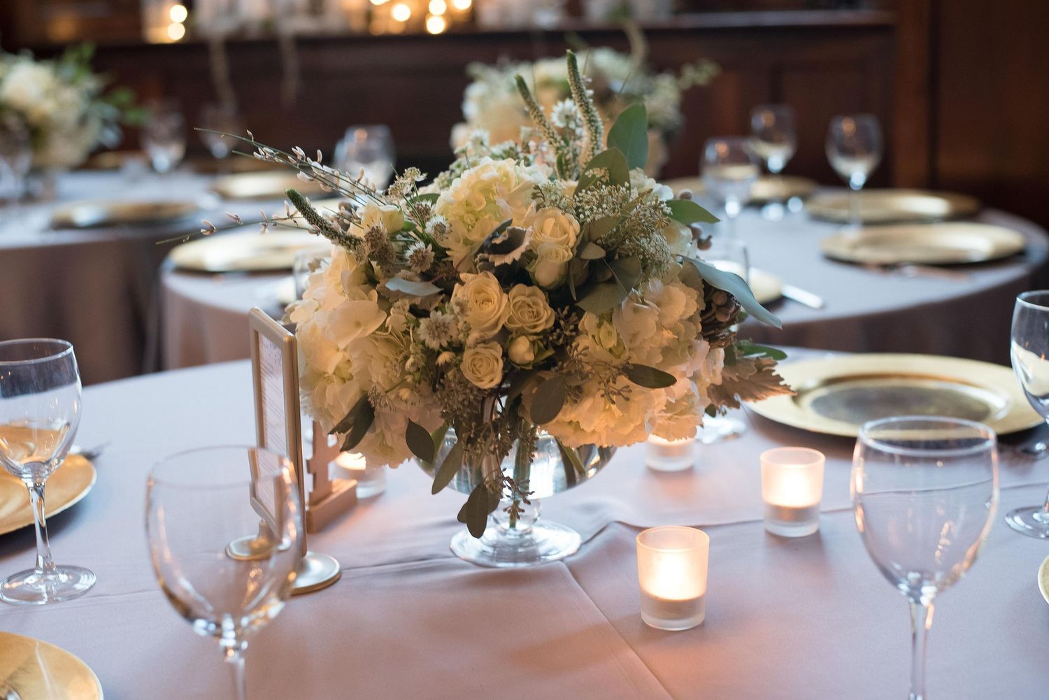 Elegant wedding centerpiece with white roses, greenery, and candles on a gray linen table with gold chargers.
