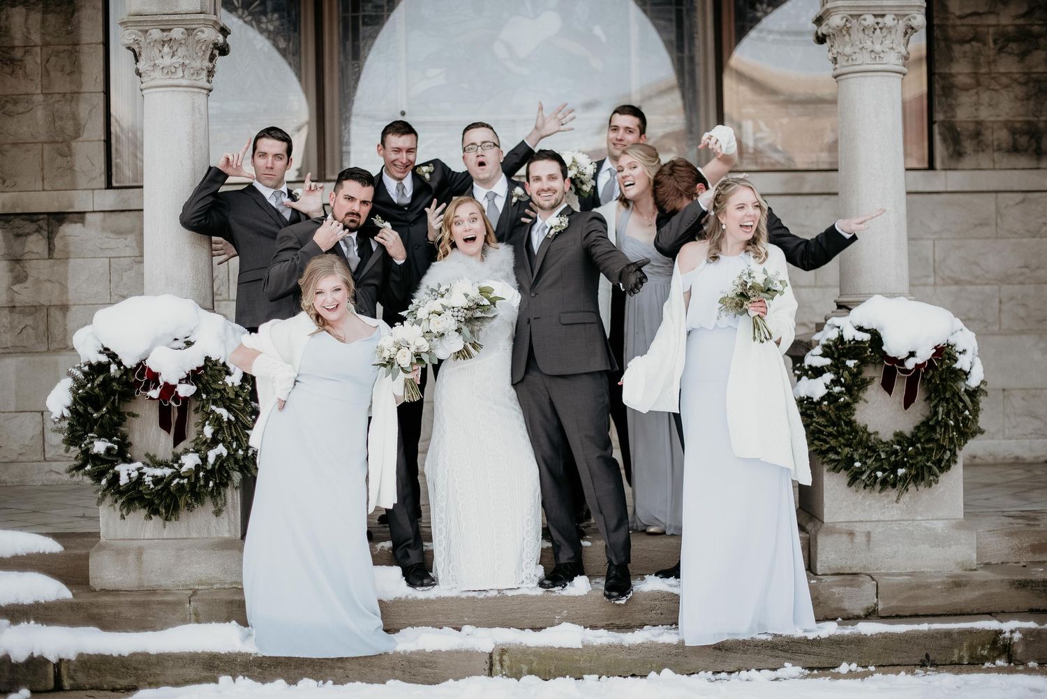 Winter wedding party poses on snowy steps, bridesmaids in silver gowns, groomsmen in gray suits, cheering outdoors.