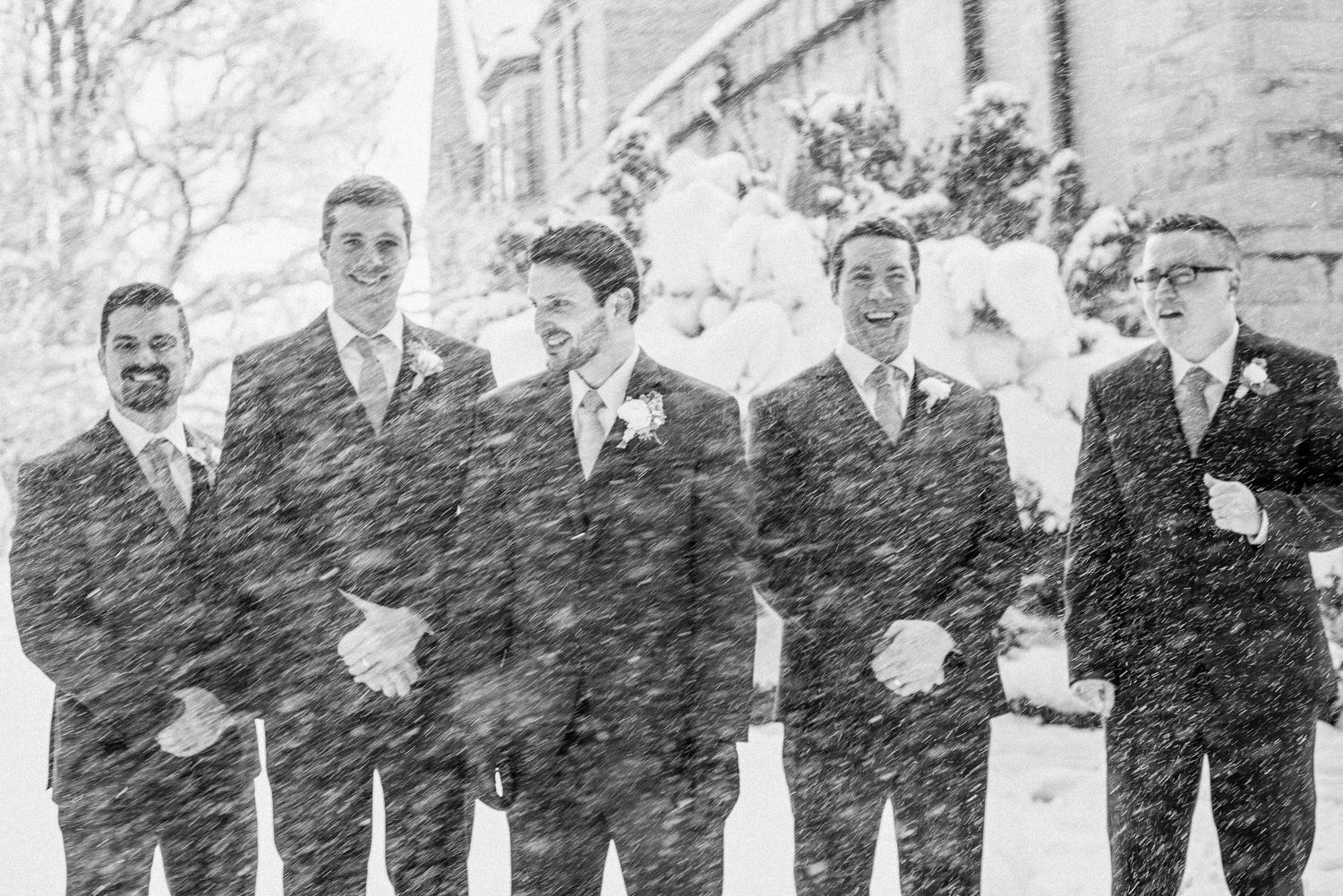 Groomsmen standing together in heavy snowfall wearing dark suits with boutonnieres at a winter wedding.
