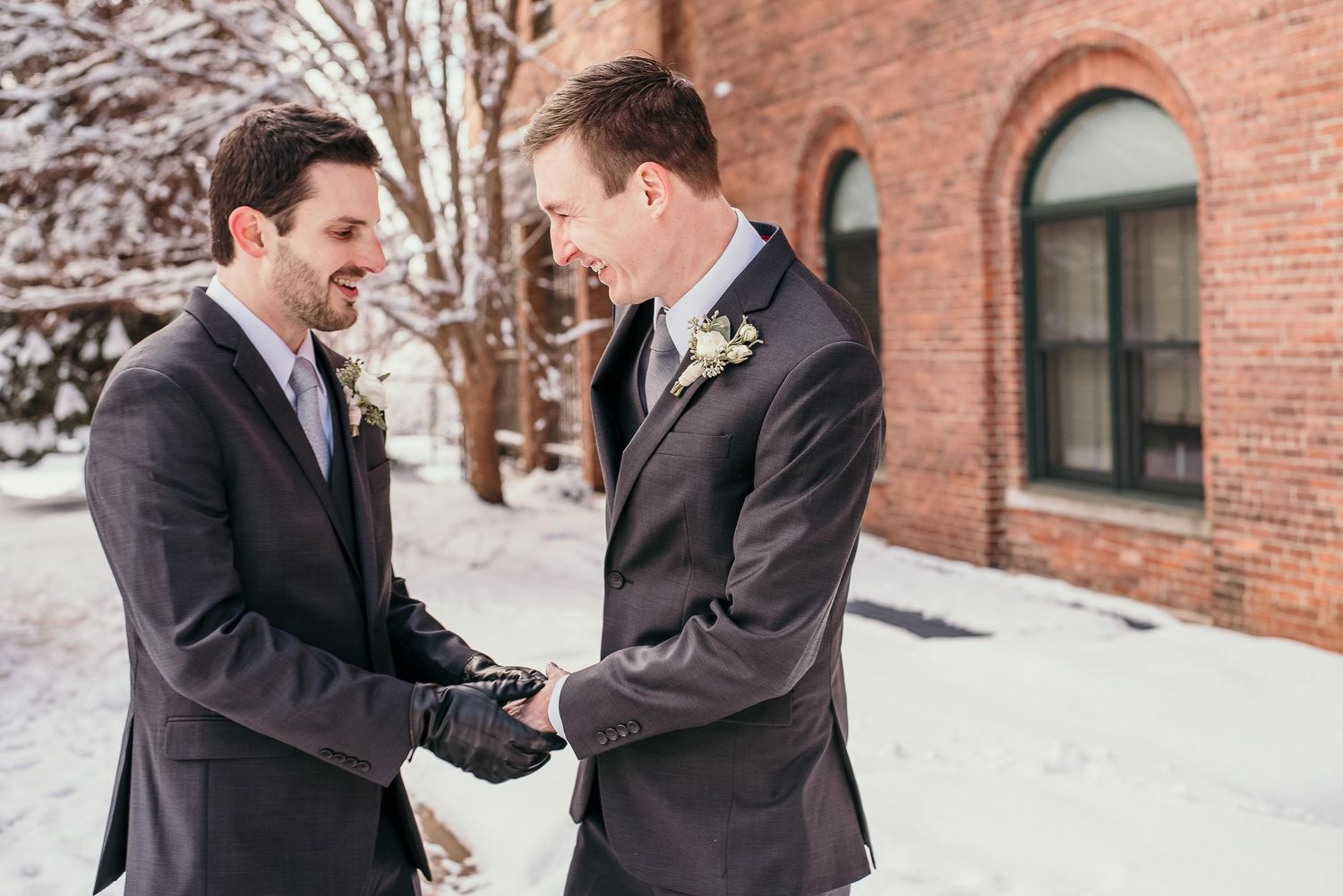 Two grooms in gray suits hold hands and smile at each other outdoors in a snowy winter wedding setting.
