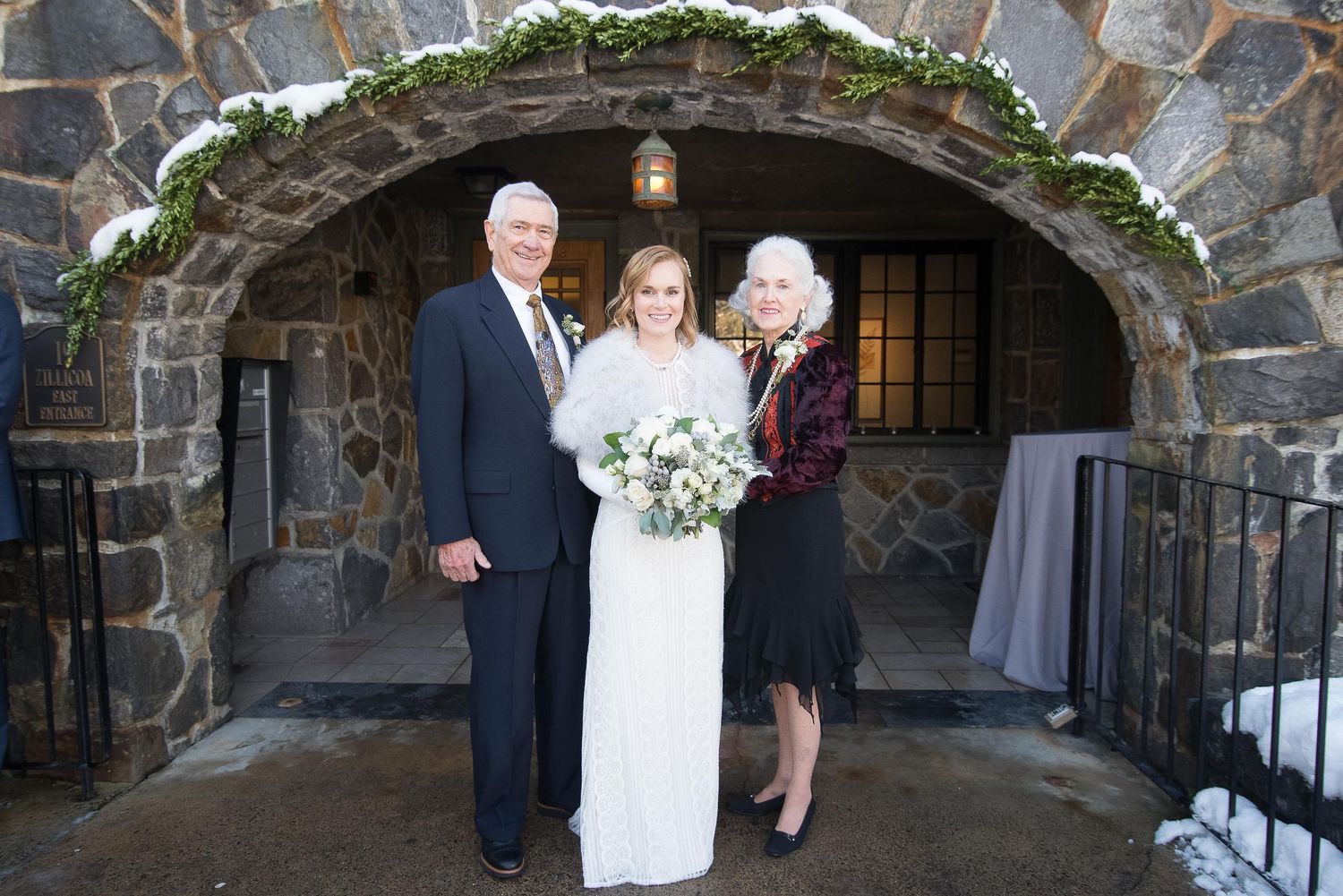 Bride in white gown poses with two guests under stone arch adorned with garland at winter wedding venue.