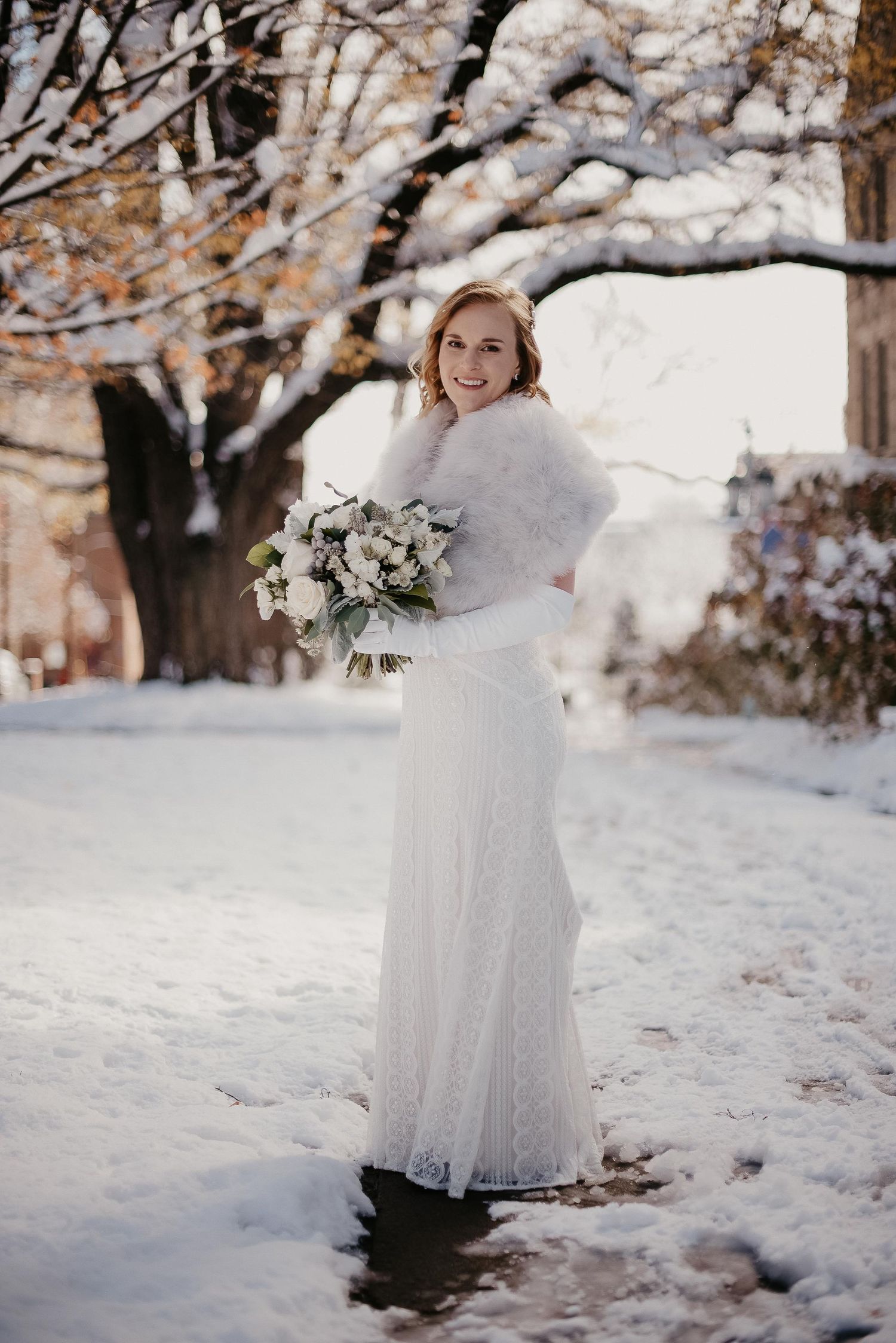 Smiling bride in white lace gown and fur wrap holds bouquet in snowy winter outdoor setting.