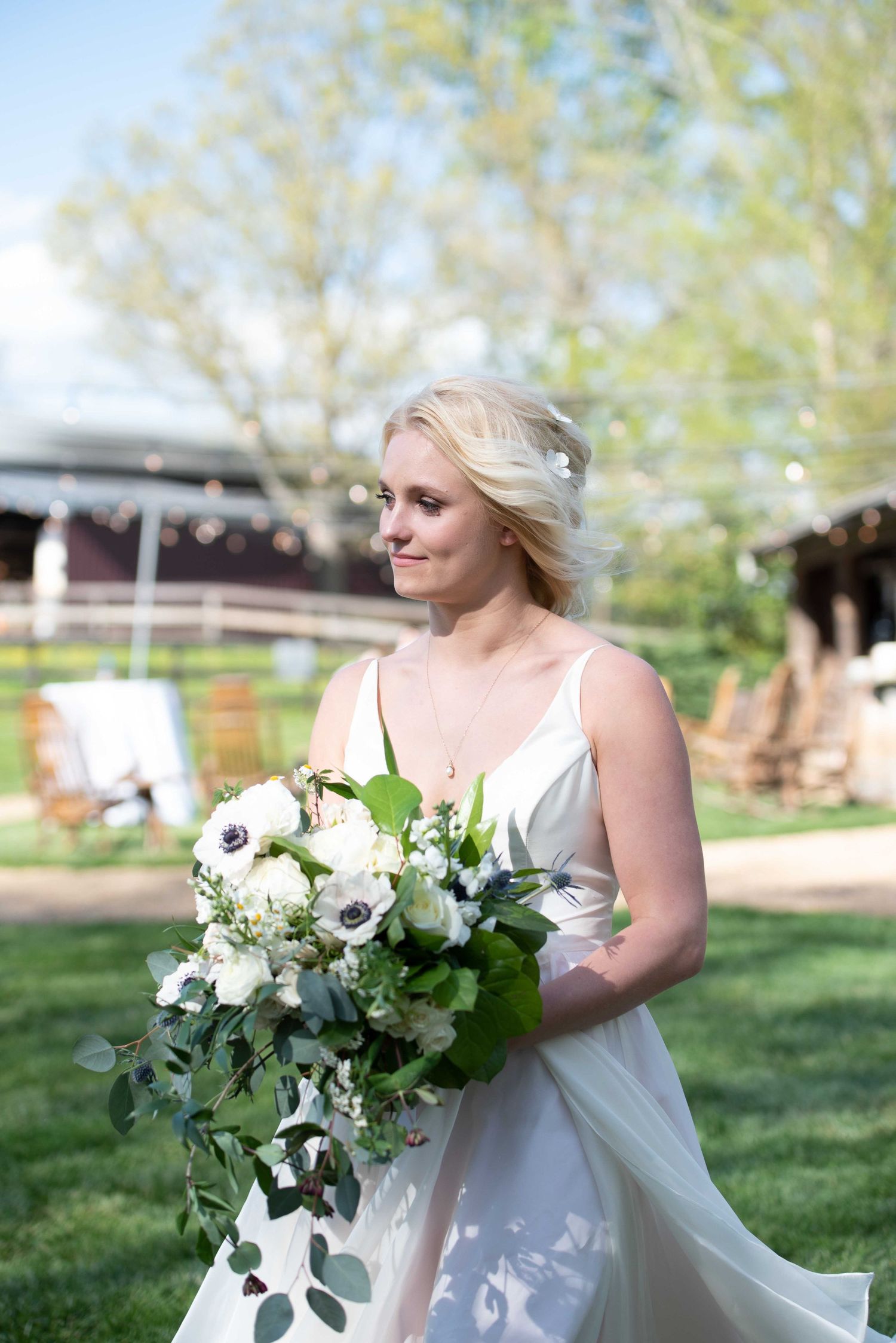 Blonde bride in white gown holds lush white and green bouquet with anemones outdoors at a rustic wedding venue.