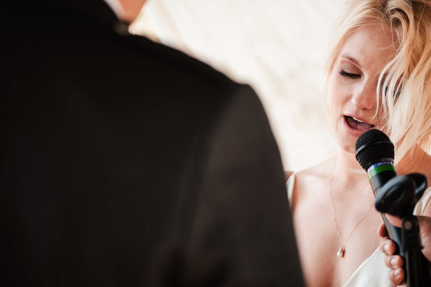 Blonde woman singing into a microphone at a wedding ceremony, wearing a white dress and delicate necklace.