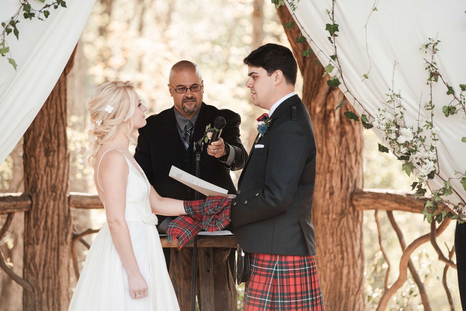 Groom in red tartan kilt exchanges vows with bride at rustic outdoor wedding ceremony with officiant.