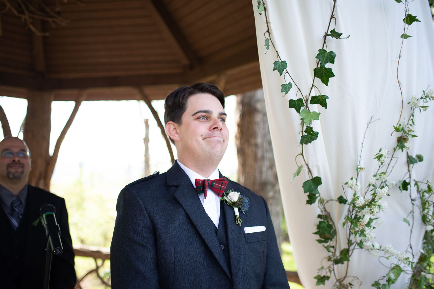 Emotional groom smiling during outdoor wedding ceremony wearing navy suit with burgundy bow tie and boutonniere.