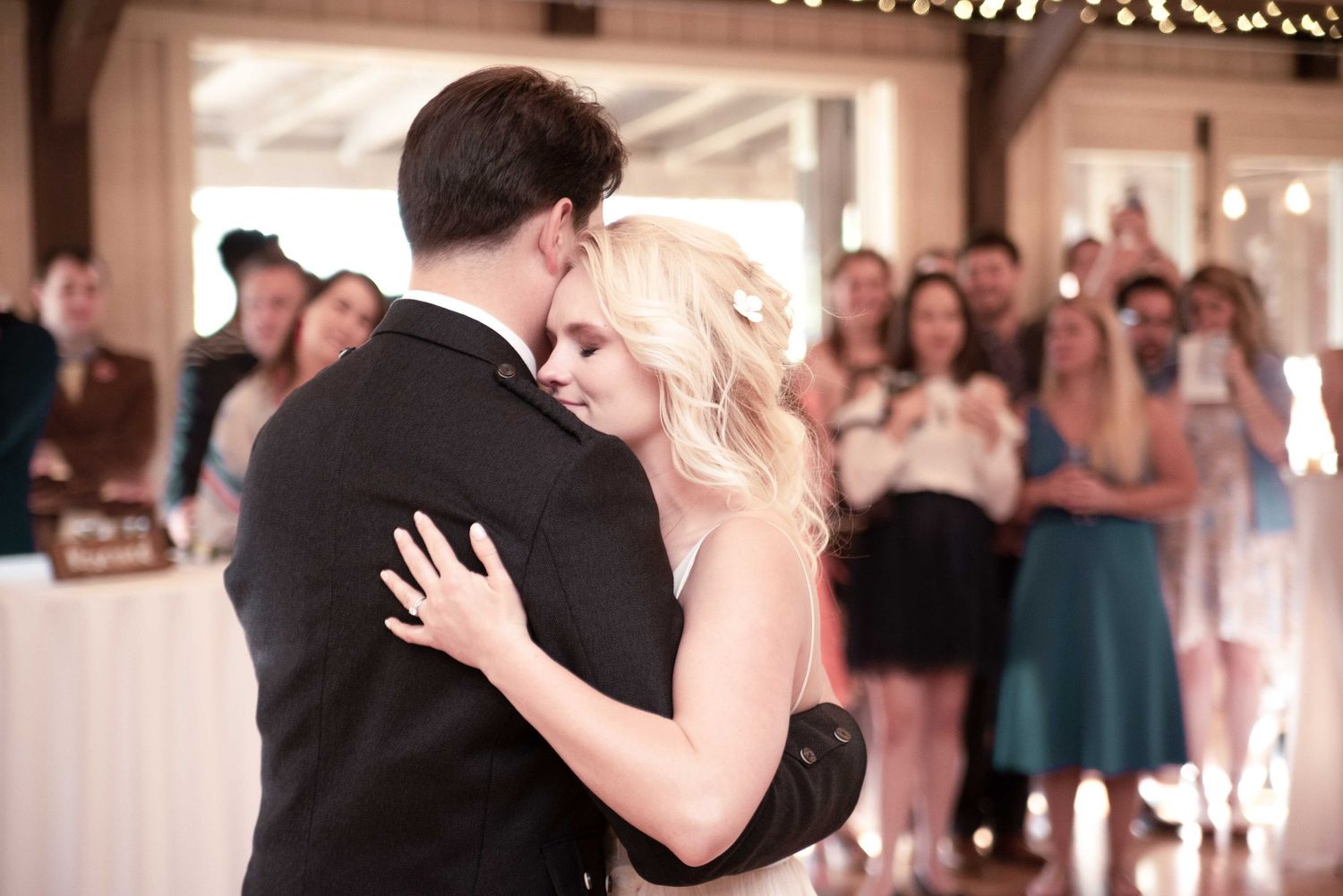 Bride and groom sharing their first dance while wedding guests watch and celebrate in a warmly lit reception venue.