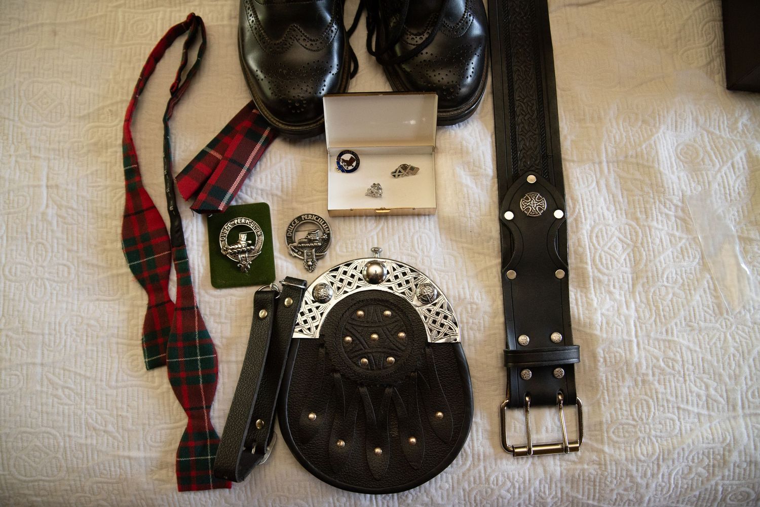 Flat lay of Scottish wedding accessories including tartan bow tie, sporran, belt, cufflinks, and black dress shoes.