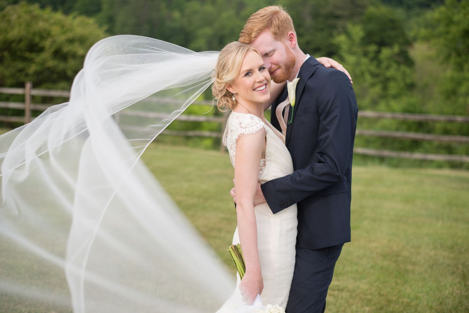 Bride and groom embrace outdoors as her flowing veil billows in the wind on wedding day.