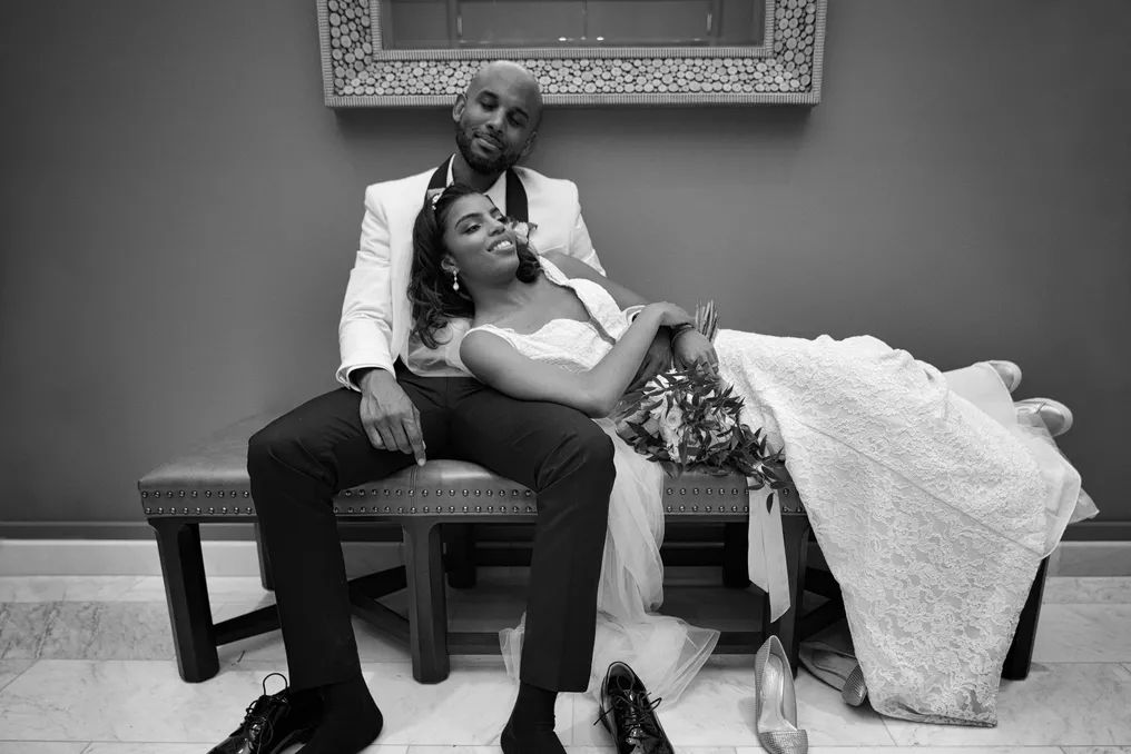 Black and white wedding photo of bride and groom sitting together on a bench in elegant attire.