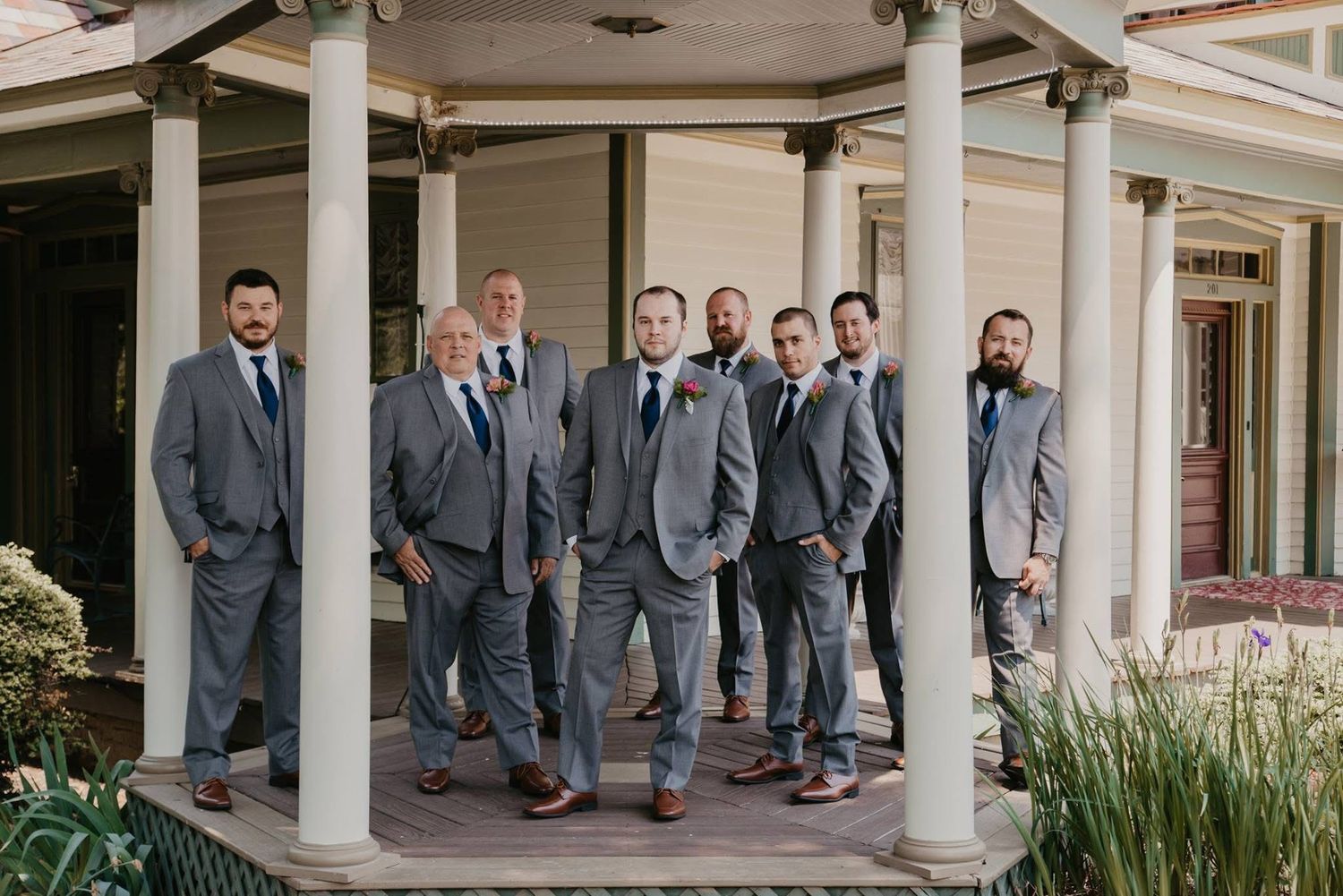 A groom and eight groomsmen in matching gray suits with navy ties pose on a elegant venue porch.