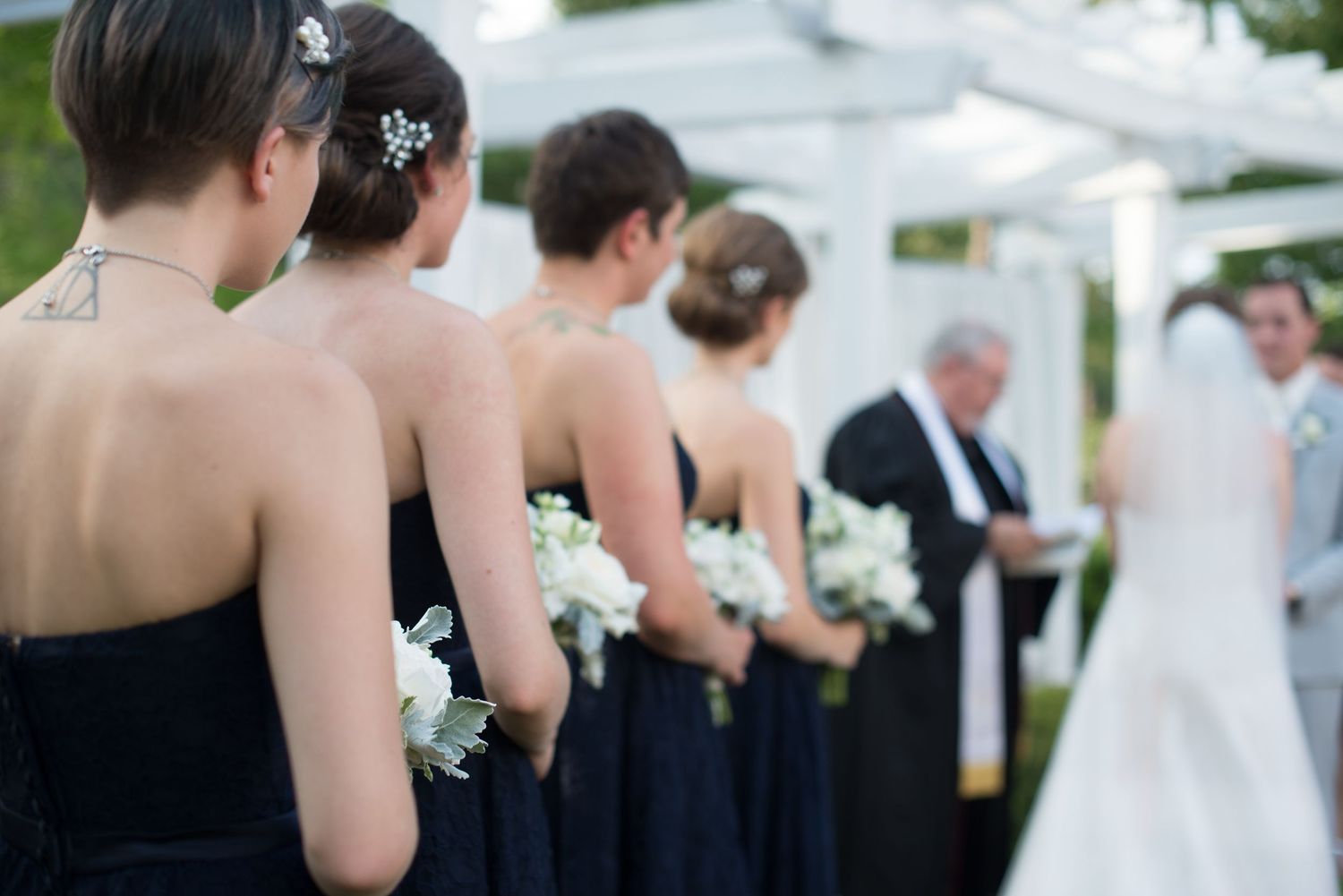 Bridesmaids in navy strapless gowns holding white bouquets stand at outdoor wedding ceremony while officiant addresses couple.