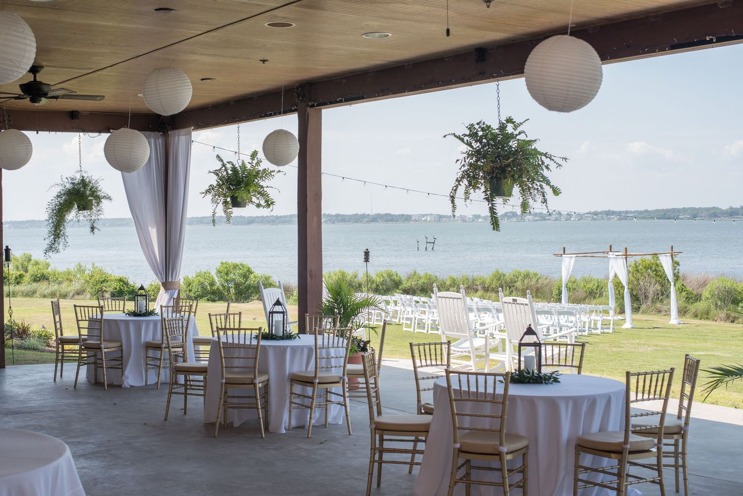 Elegant waterfront wedding venue with white draped tables, hanging plants, and outdoor ceremony area.