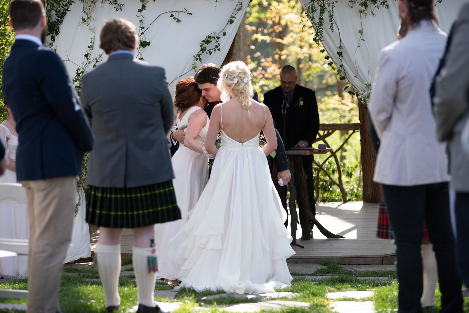 Bride in backless white gown shares first kiss with groom at outdoor wedding altar.
