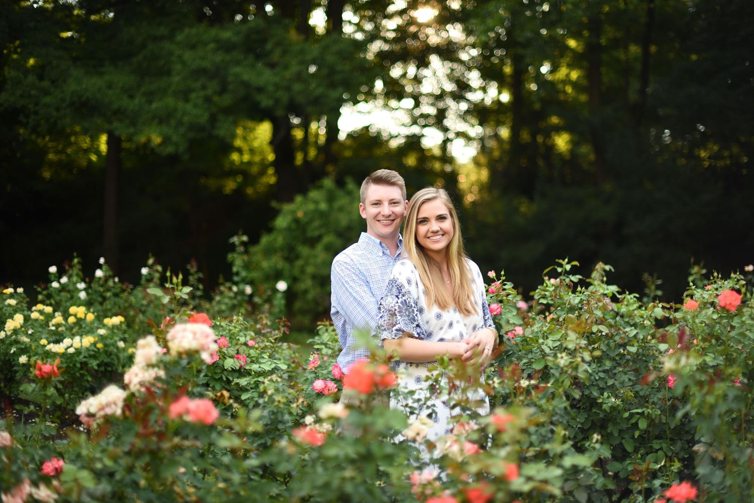 Couple embracing in a colorful rose garden at golden hour with lush green trees in the background.