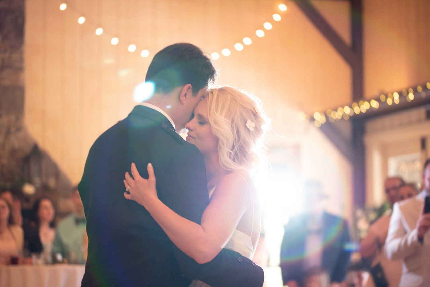 Bride and groom share romantic first dance kiss under warm string lights at wedding reception.