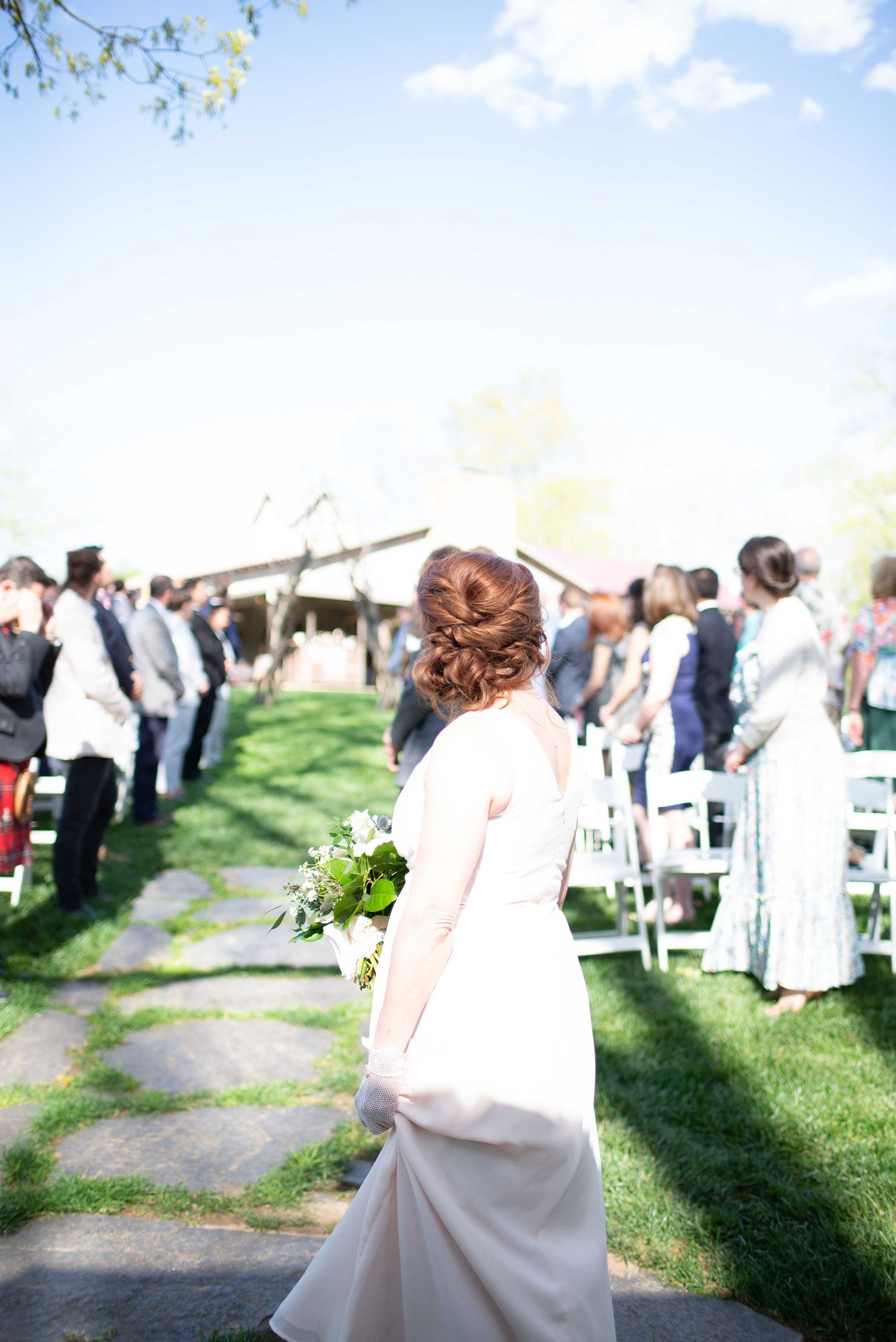 Bride in white gown walks down outdoor stone aisle toward wedding guests seated on white chairs on a sunny day.