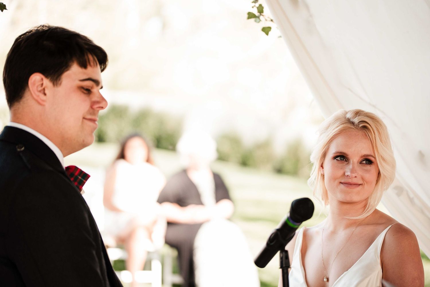 Bride and groom exchanging vows at outdoor wedding ceremony under white tent with microphone stand.