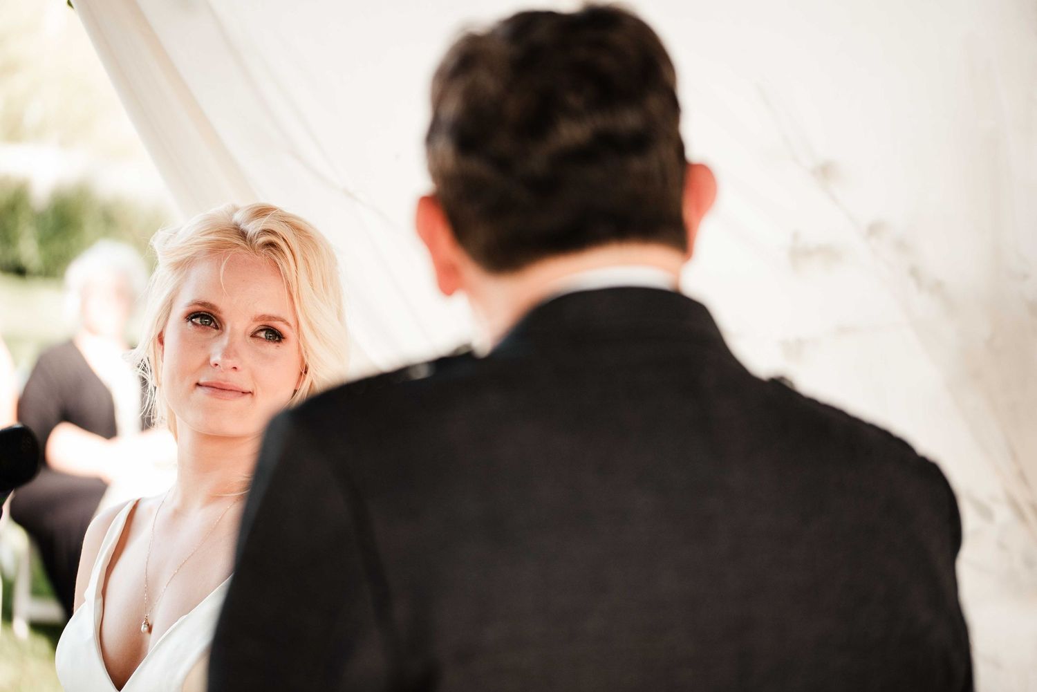 Bride gazing lovingly at groom during outdoor wedding ceremony under white tent.