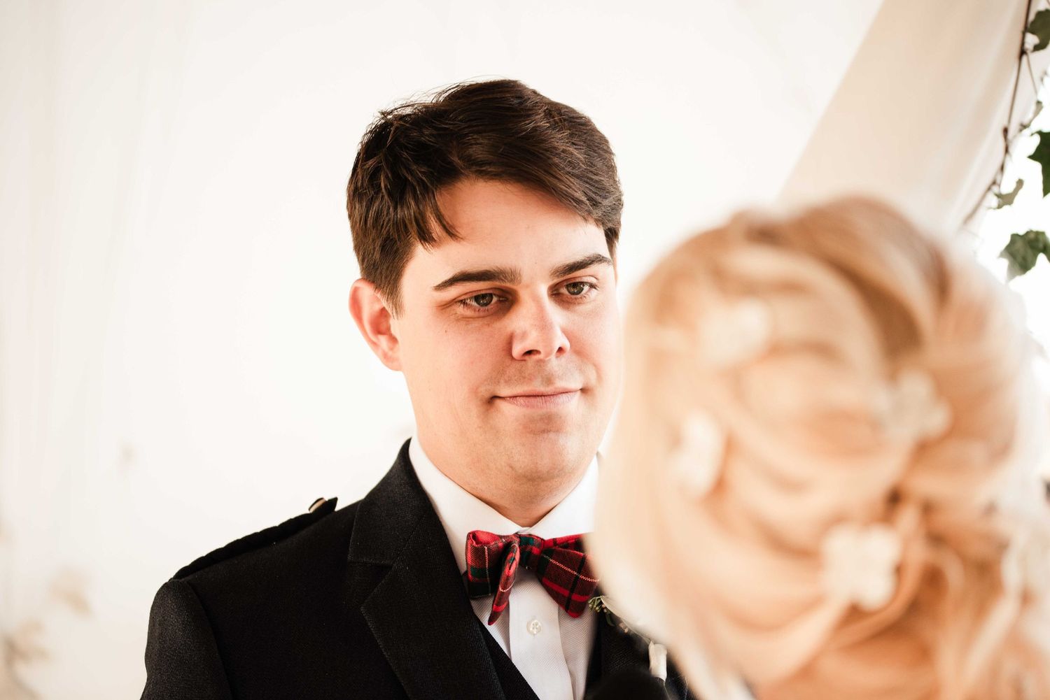 Groom in black tuxedo and burgundy bow tie gazes lovingly at blonde bride during wedding ceremony.