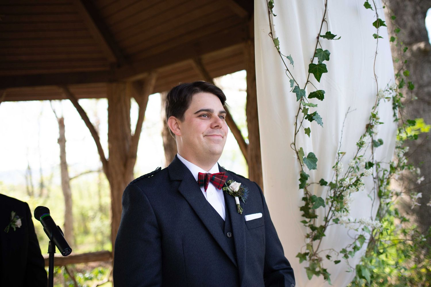 Smiling groom in navy suit and burgundy bow tie standing at outdoor wedding ceremony altar with ivy decor.