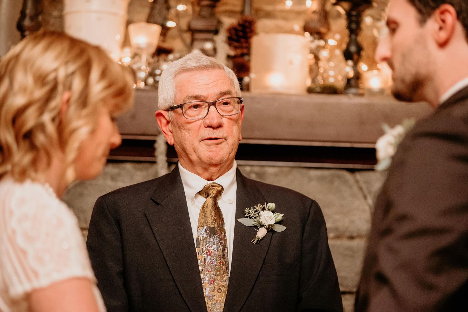 Elderly man in suit with boutonniere officiating a wedding ceremony between a bride and groom indoors.