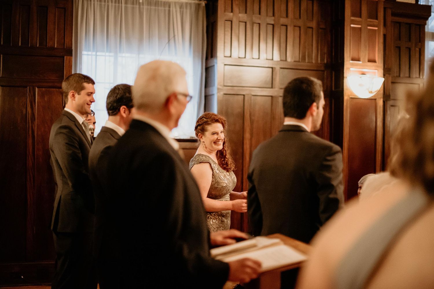 Bride smiling joyfully during an intimate wedding ceremony in an elegant wood-paneled room with guests nearby.