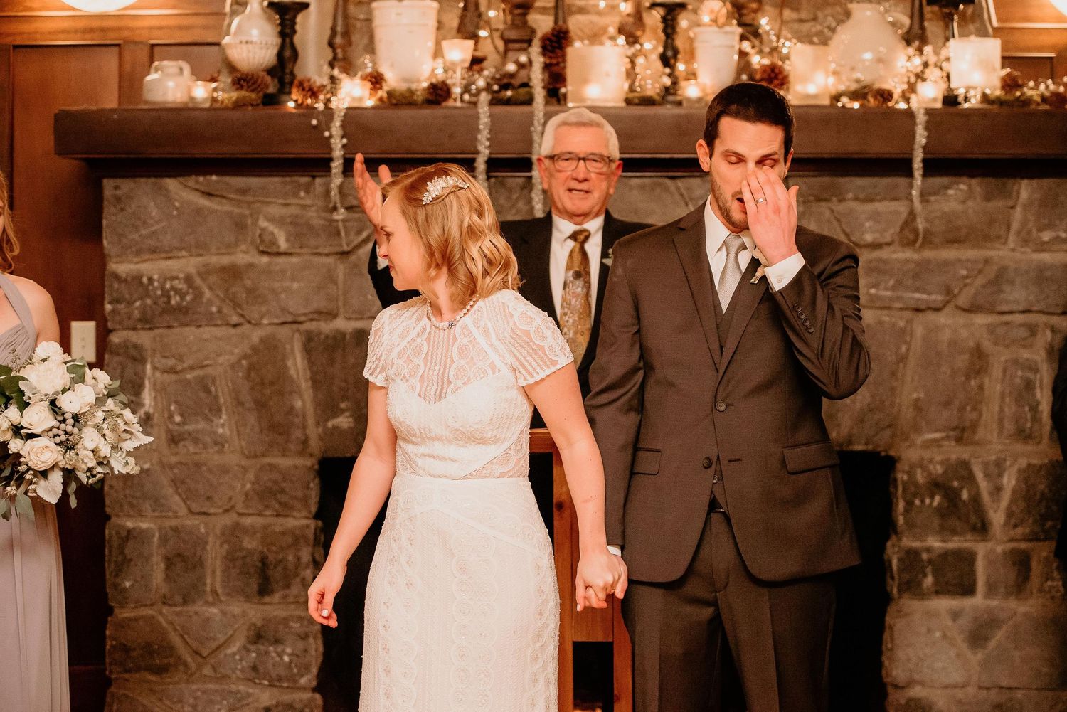 Emotional groom wiping tears during indoor wedding ceremony by stone fireplace with officiant and bride holding hands.