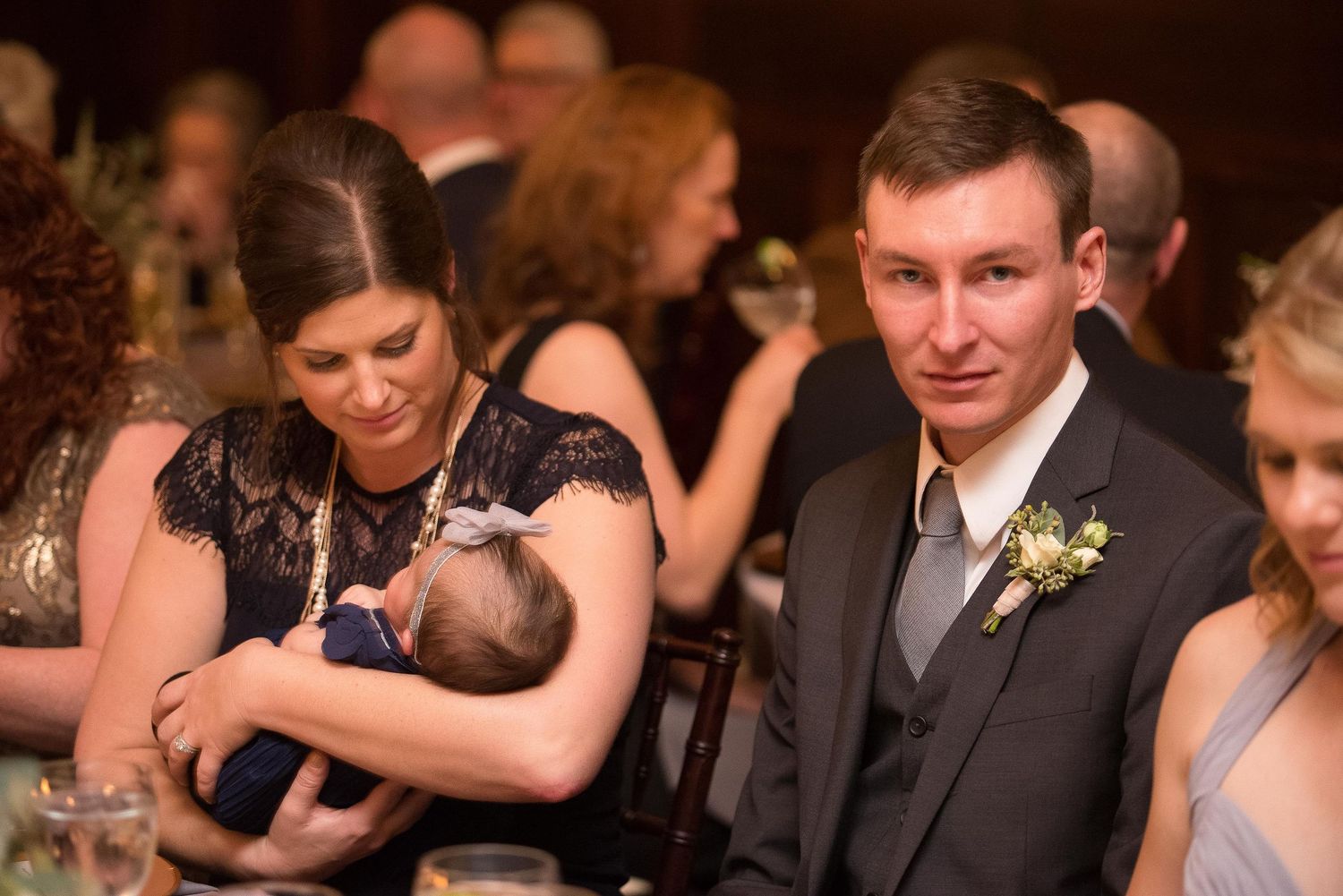A woman in a navy lace dress holds a newborn baby at a wedding reception while a man in a gray suit sits beside her.