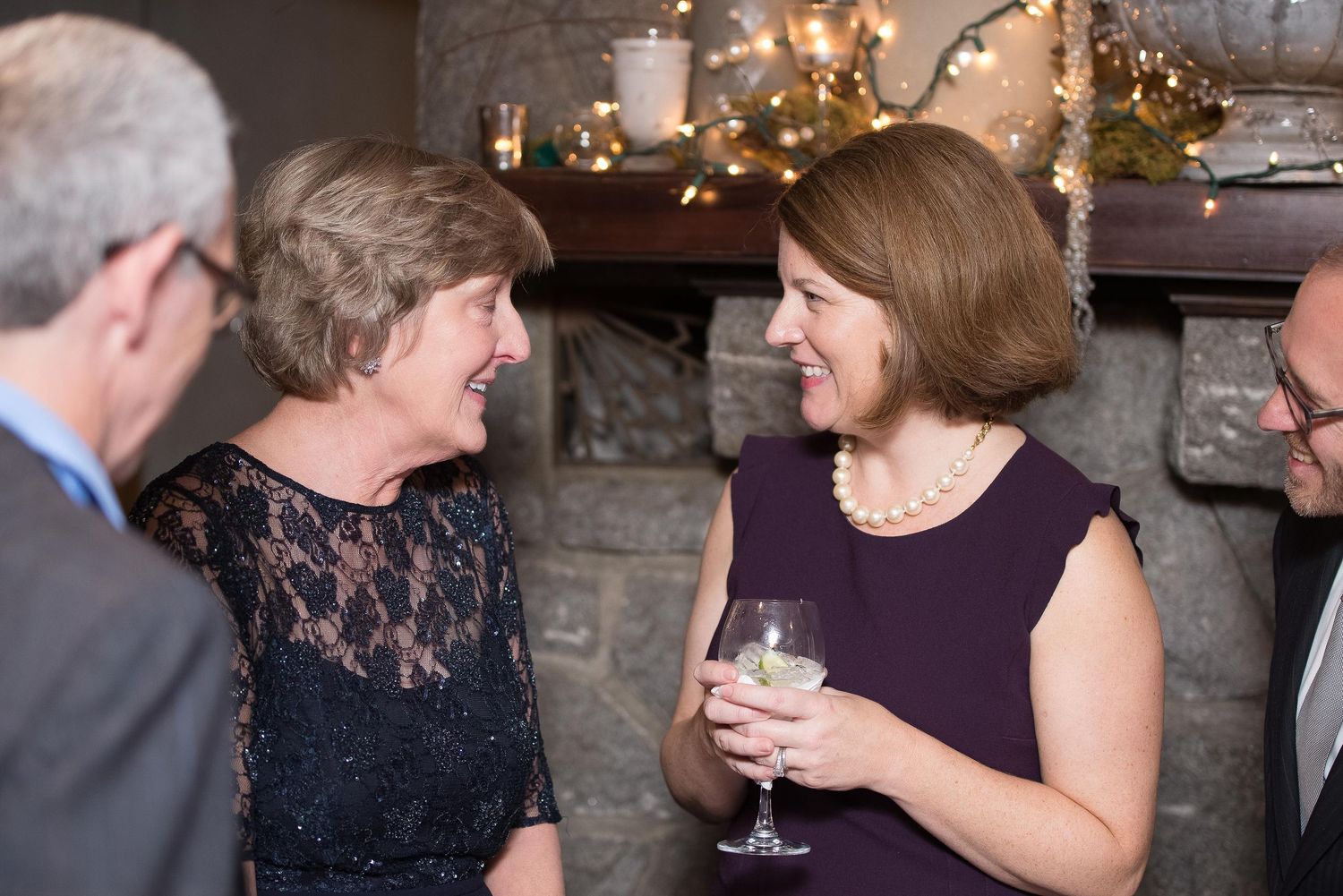 Two women share a laugh at an elegant holiday party, one holding a wine glass, near a decorated fireplace.