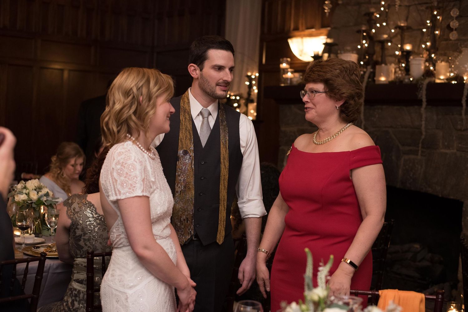 Bride in white lace dress and groom in vest chat with woman in red off-shoulder dress at elegant wedding reception.