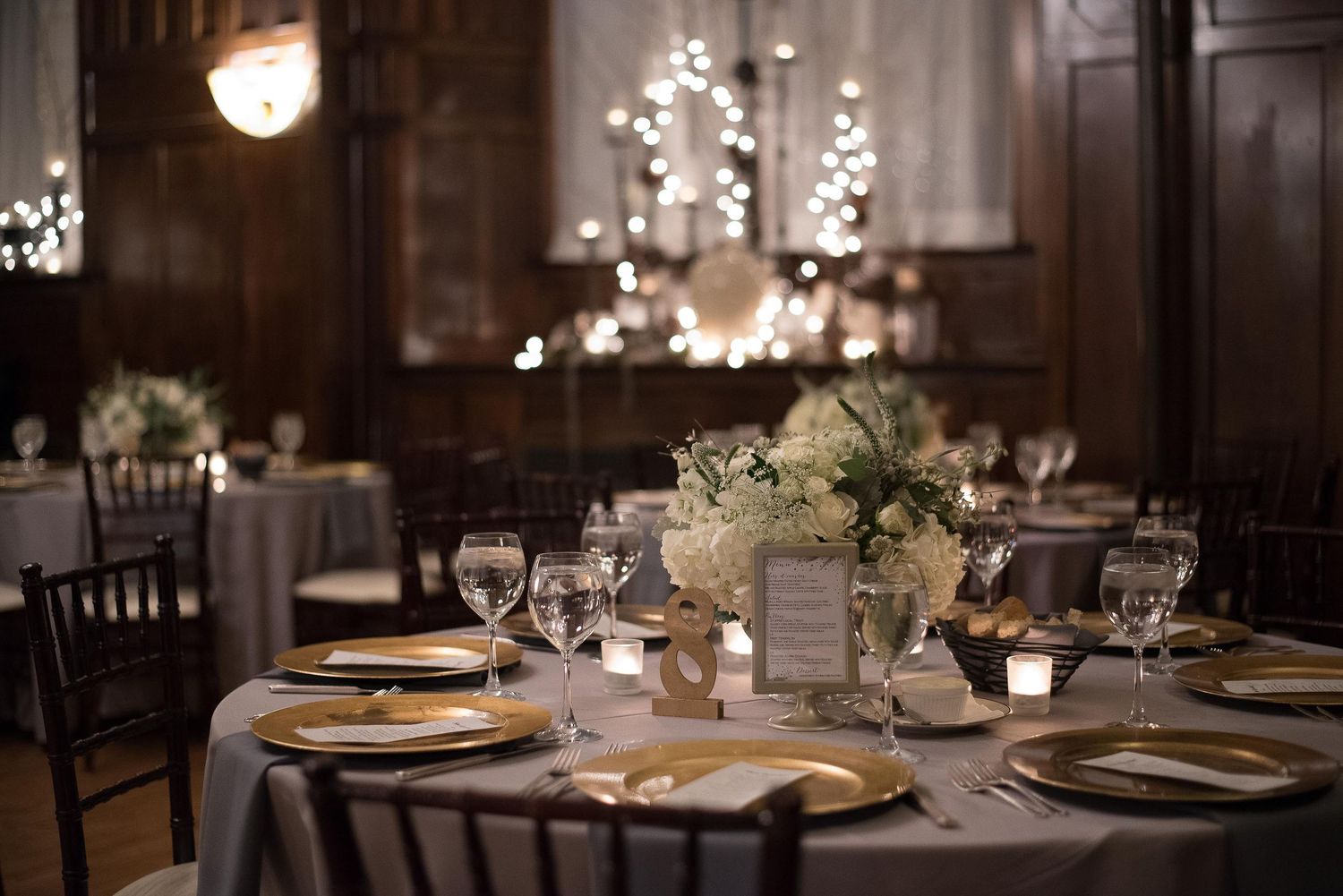 Elegant wedding reception hall with candlelit tables, gold chargers, white floral centerpieces, and warm bokeh lights in background.