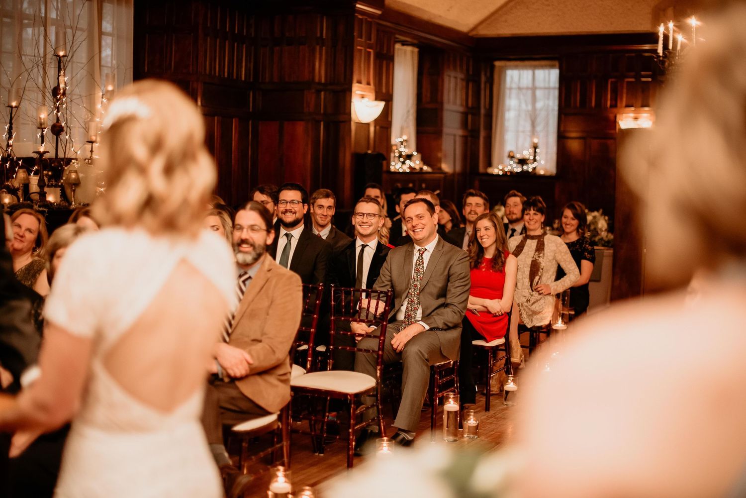 Wedding guests smile and watch ceremony in elegant wood-paneled room as bride stands at altar.