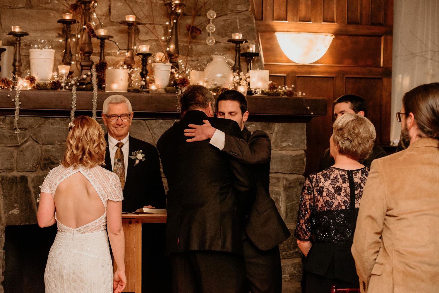 Two grooms share an emotional hug at wedding altar near stone fireplace with candles and guests watching.