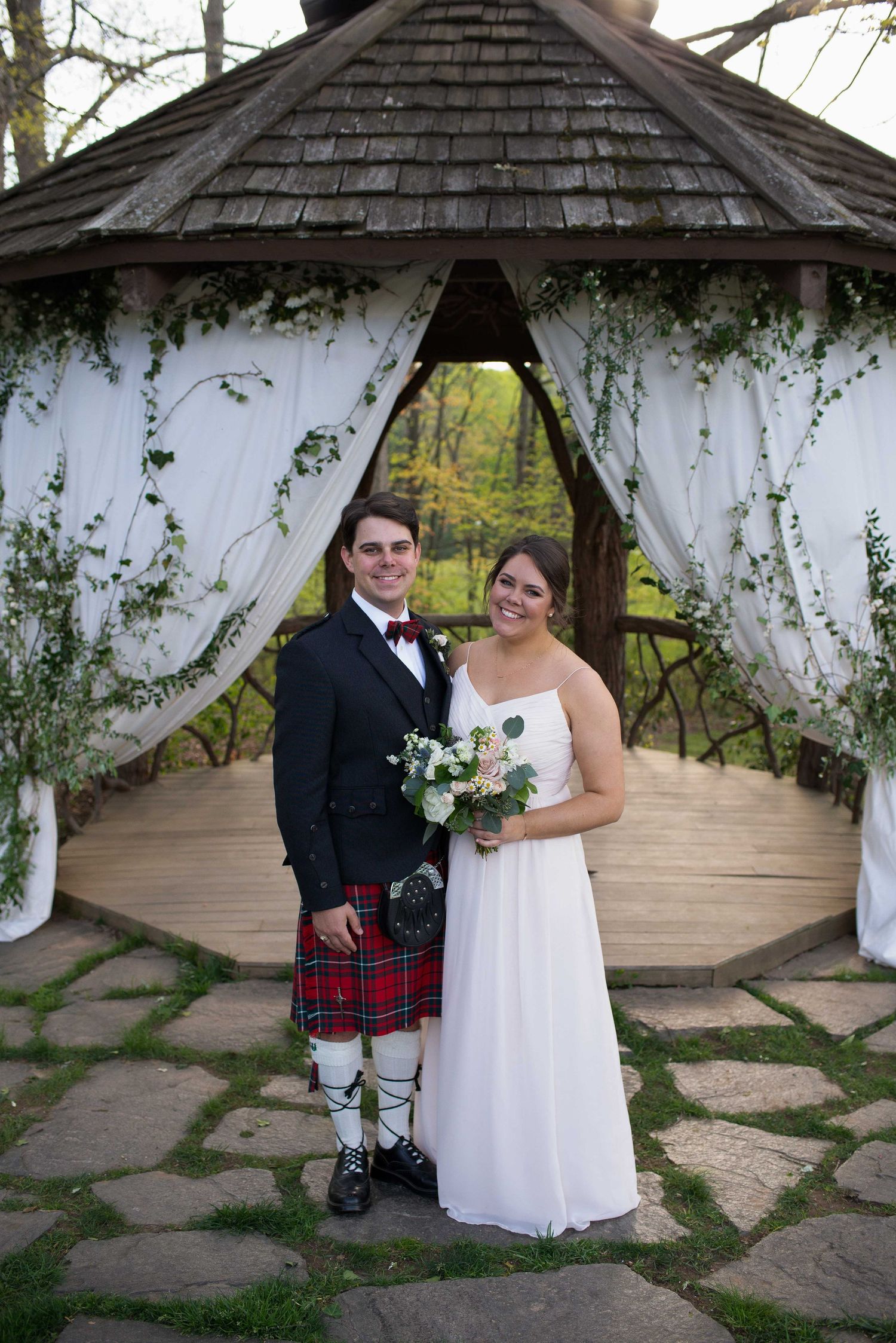 Bride in white gown and groom in black jacket with red tartan kilt pose under a decorated gazebo on their wedding day.